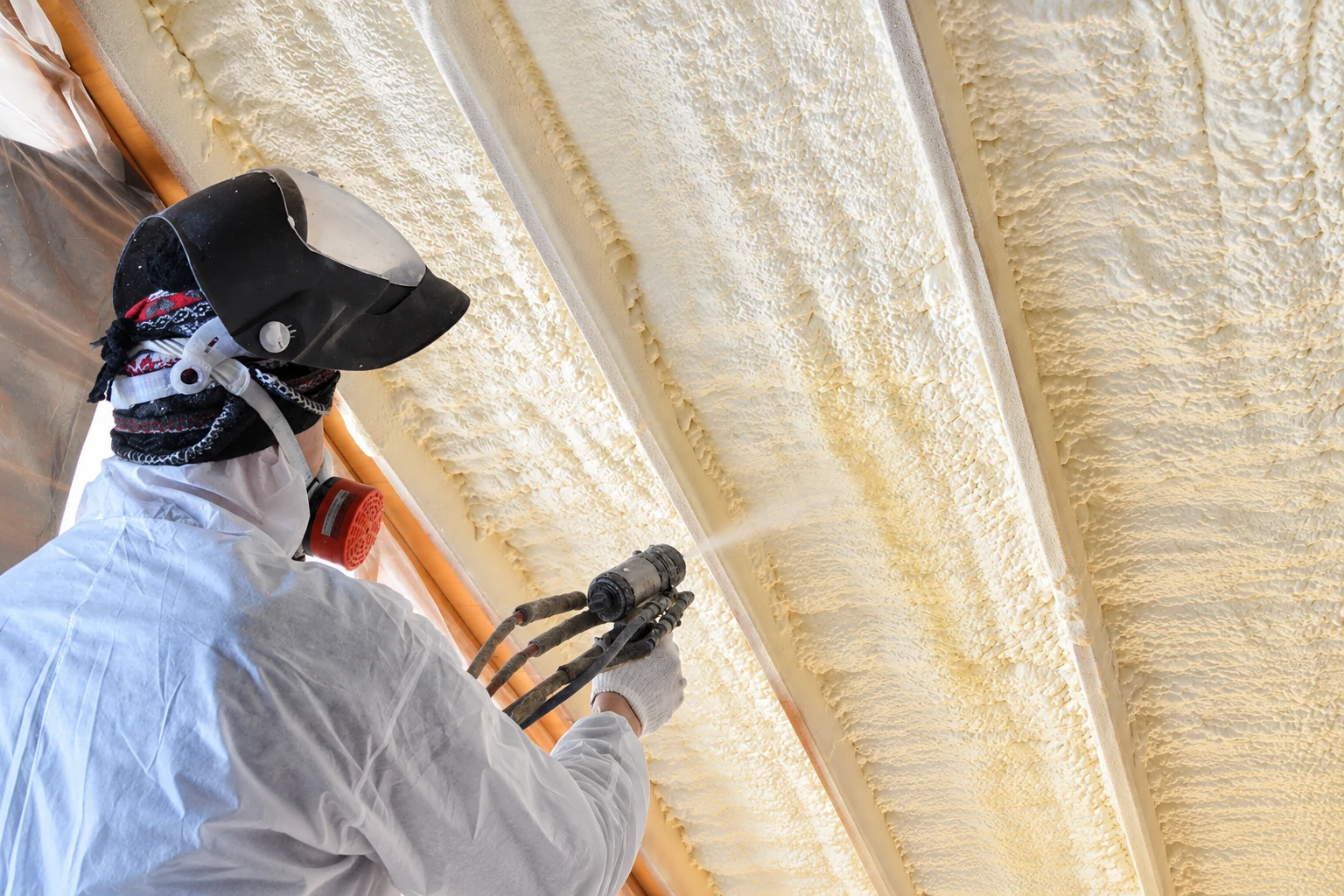 A worker wearing protective gear aims a spray-foam gun at the insulated rafters of an attic.