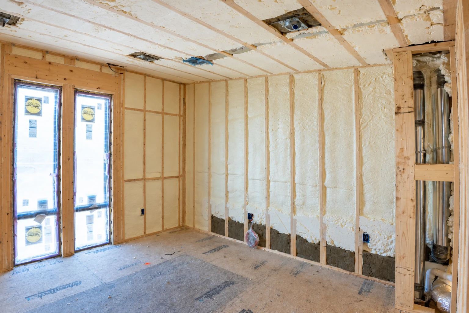 Cream-colored spray foam fills wall and ceiling cavities in an unfinished room with exposed framing and tall windows.
