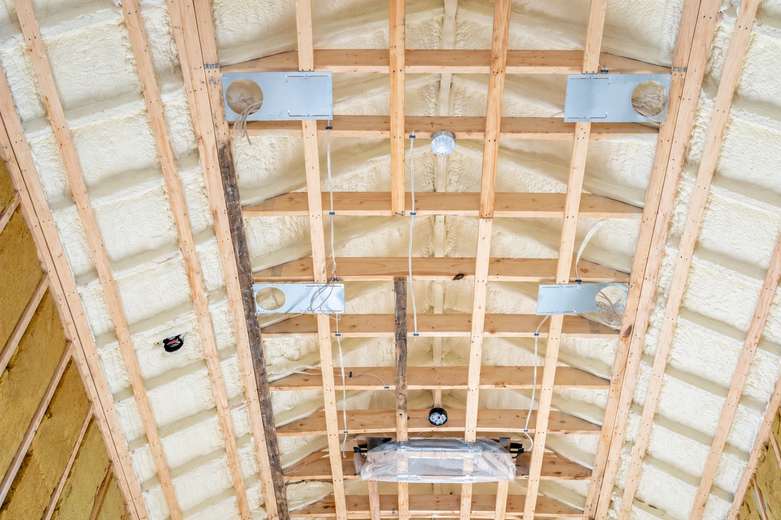 White spray foam coats the roof framing around exposed beams, wiring, and metal fixtures.