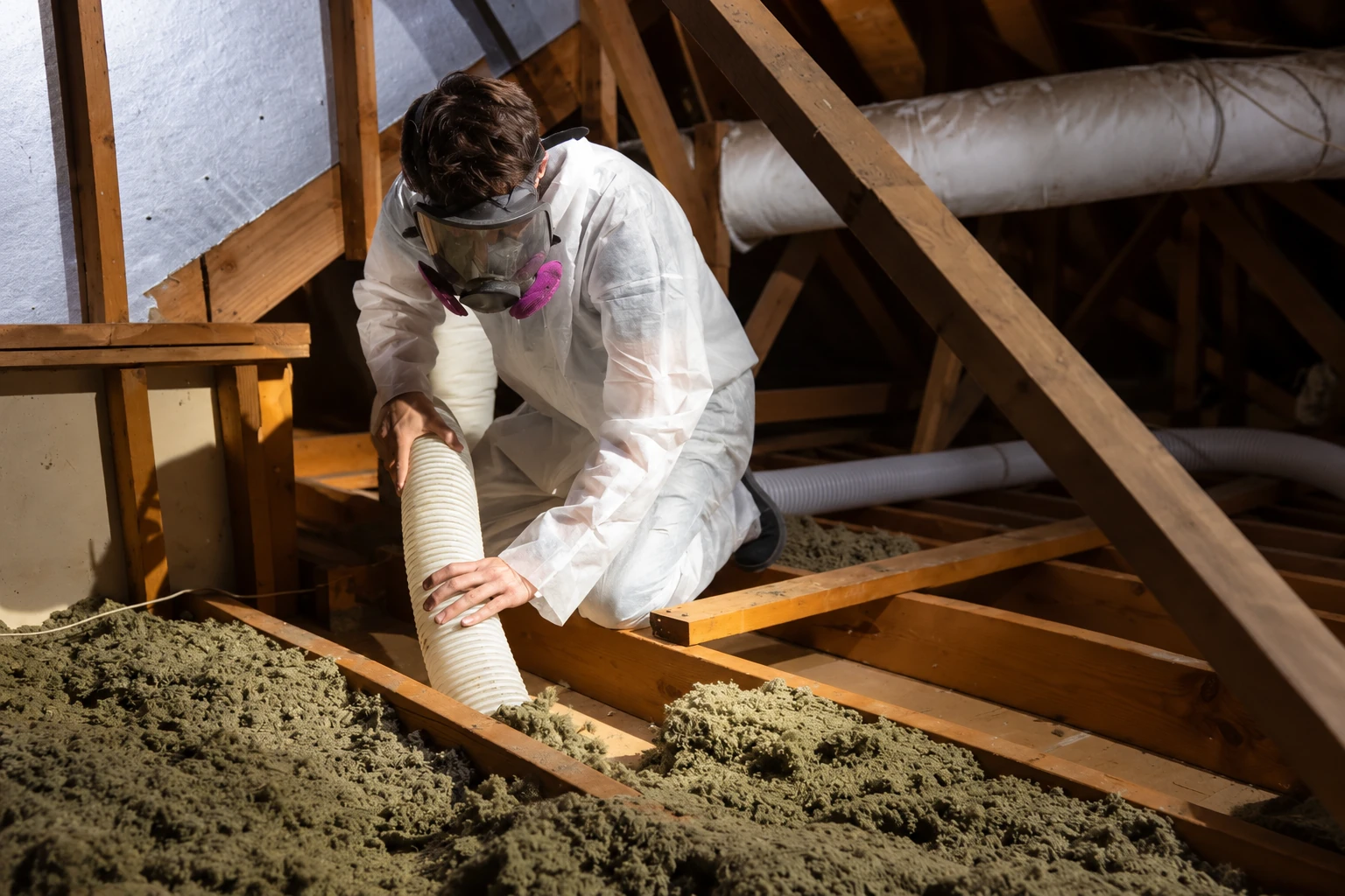 A worker in protective coveralls and a respirator guides a large hose through loose attic insulation between exposed wooden joists.
