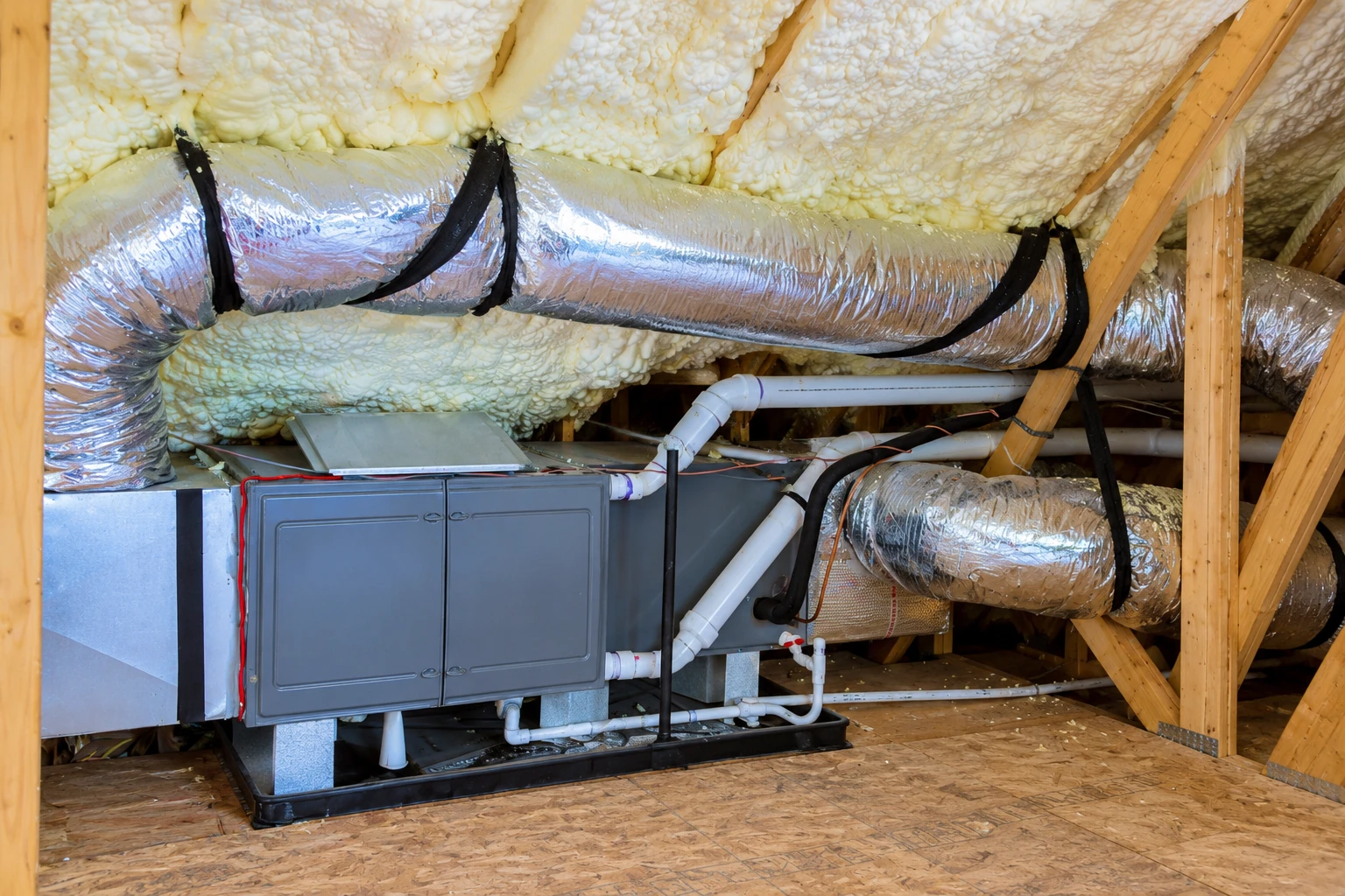 Foam insulation coats the attic roof around a large silver flexible duct and a gray HVAC unit.