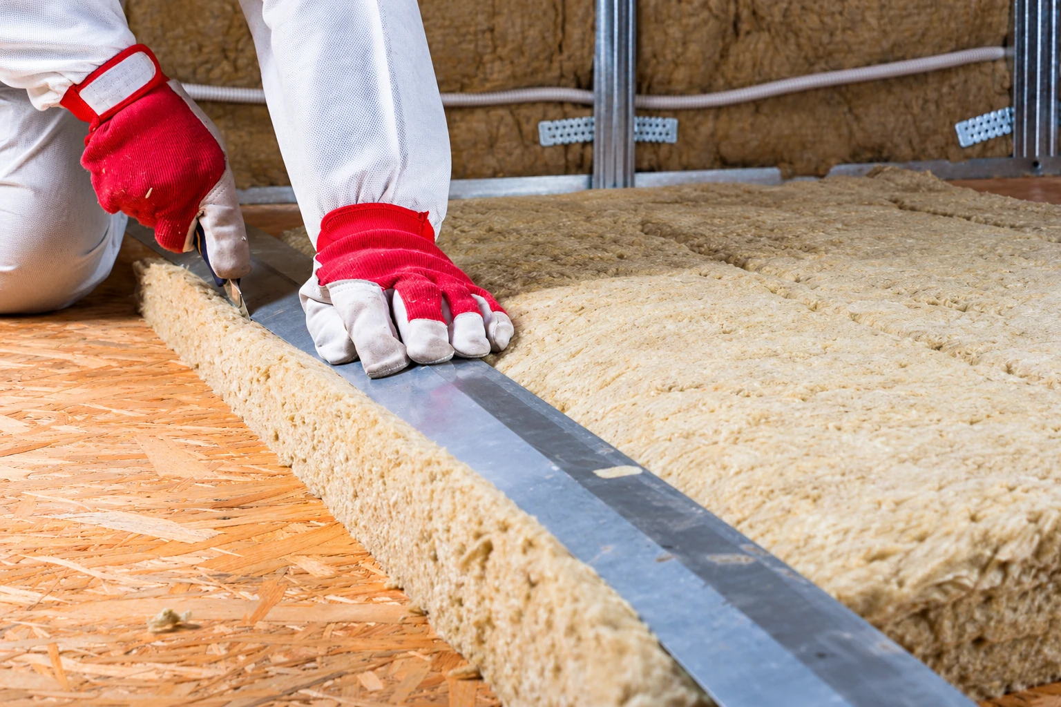 A worker in red-and-white gloves cuts a thick insulation batt beside a metal straightedge on an unfinished floor.