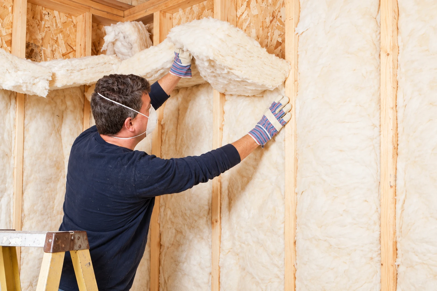 A masked worker in striped gloves fits fluffy fiberglass batts between the wooden wall studs.