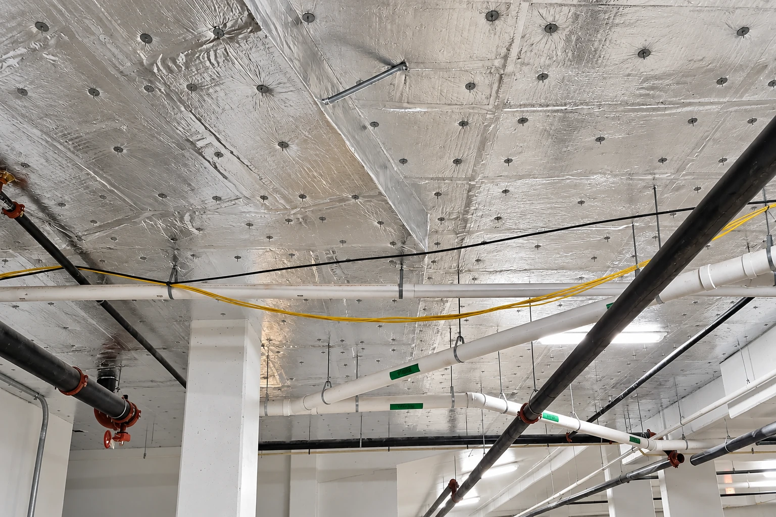 Reflective vapor barrier covers a commercial ceiling, secured with round fasteners around exposed pipes and cables.
